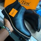 A hand holding a pair of black G-Ten leather boots with a yellow tag attached. The boots are shown from the side and have a chunky sole and a pointed toe. The brand's logo is visible on the tongue.