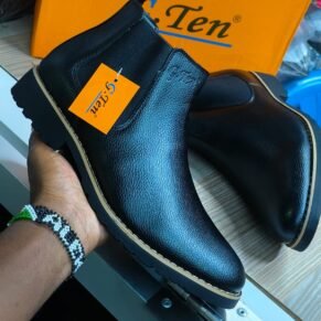 A hand holding a pair of black G-Ten leather boots with a yellow tag attached. The boots are shown from the side and have a chunky sole and a pointed toe. The brand's logo is visible on the tongue.