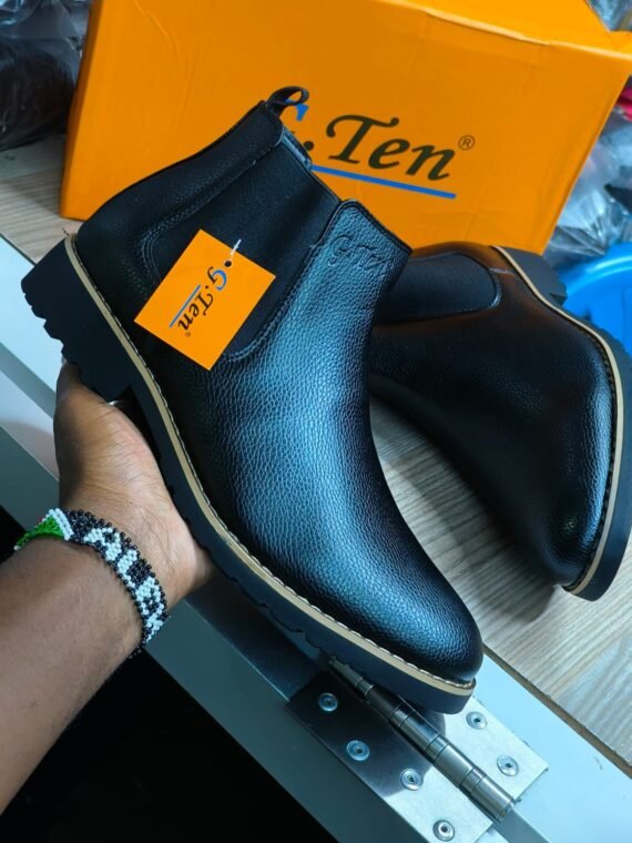 A hand holding a pair of black G-Ten leather boots with a yellow tag attached. The boots are shown from the side and have a chunky sole and a pointed toe. The brand's logo is visible on the tongue.