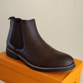 A high-quality, stylish Chelsea boot in a dark brown color, displayed on a bright orange shoebox. The boot features a classic Chelsea design with a contrasting brown leather upper and a contrasting black rubber outsole.